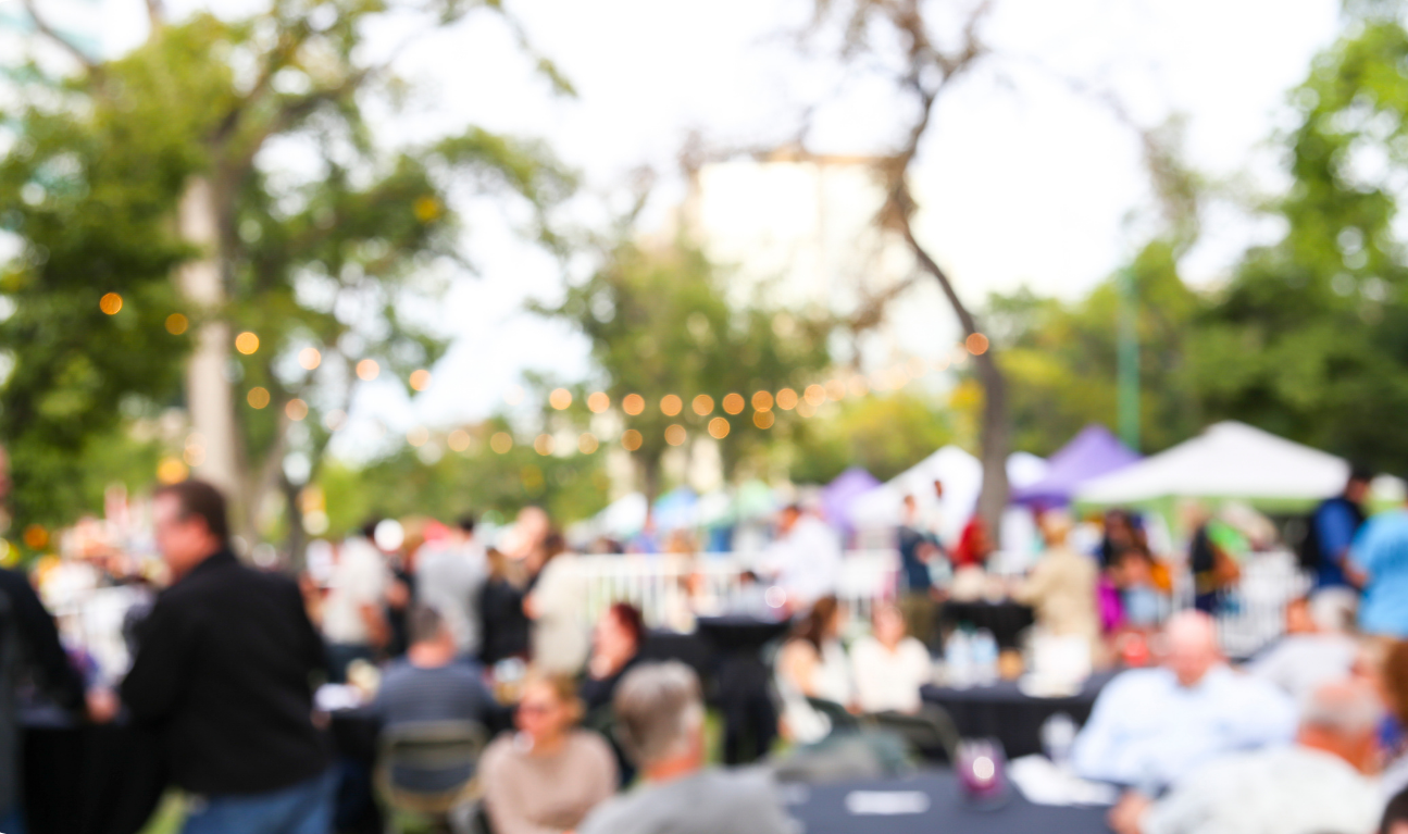 Unscharfe Aufnahme eines lebhaften Outdoor-Festivals mit Menschen an Tischen, Marktständen im Hintergrund und Lichterketten zwischen den Bäumen.